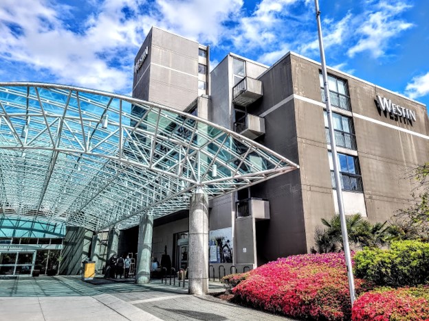 The exterior of a Westin hotel featuring a glass canopy entrance, modern architecture, and vibrant landscaping under a bright blue sky.