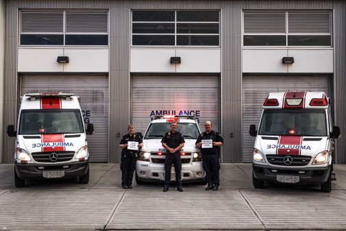 Three paramedics standing in front of two ambulances and an emergency vehicle, parked outside a modern ambulance station with garage doors.