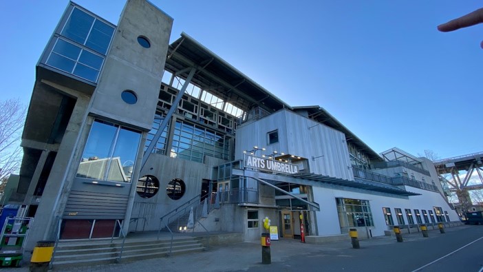 Modern arts building labeled "Arts Umbrella," featuring a mix of industrial and contemporary architectural elements with large glass windows and a concrete facade.