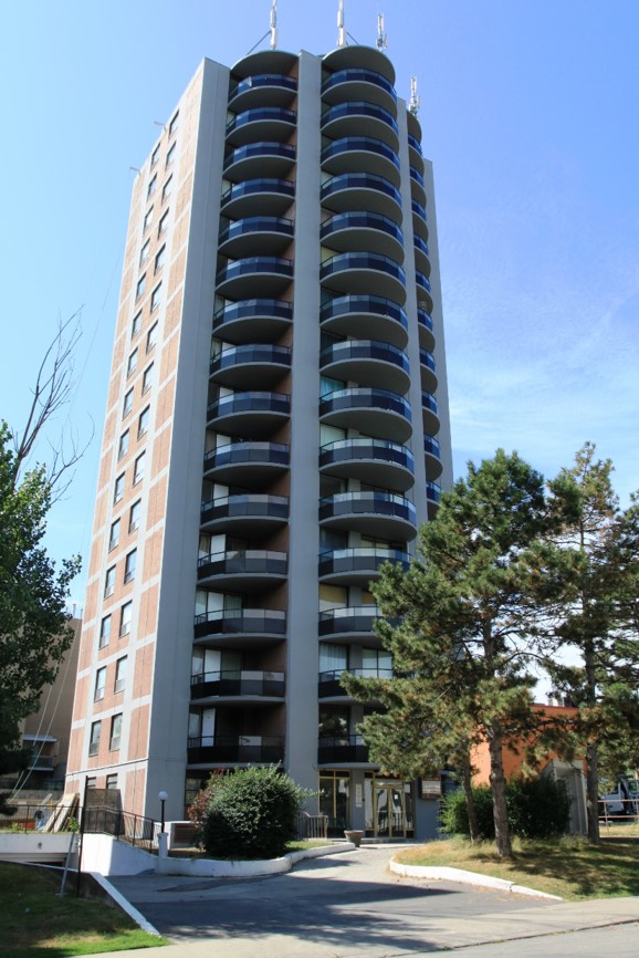 High-rise residential building with rounded balconies, surrounded by trees and a driveway leading to the entrance.