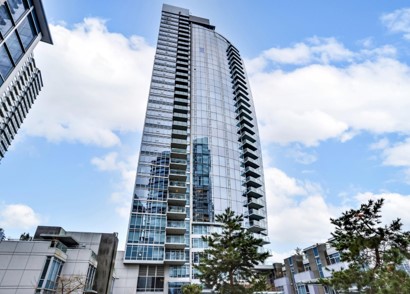 Modern glass high-rise residential building with balconies, surrounded by smaller buildings and trees under a partly cloudy sky.